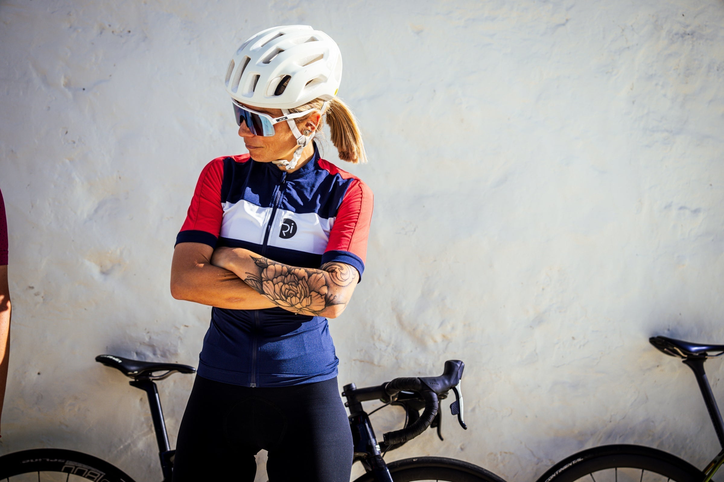 A person wearing a cycling jersey with a helmet and sunglasses, standing next to a bicycle against a light-colored wall