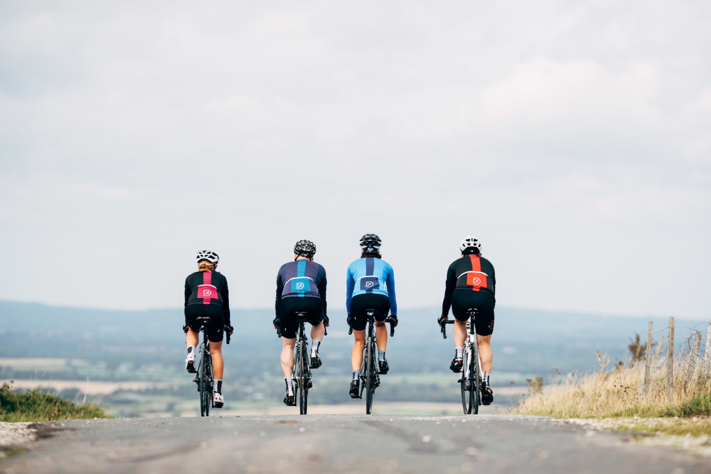 Four cyclists ride together along an open countryside road with rolling hills in the distance