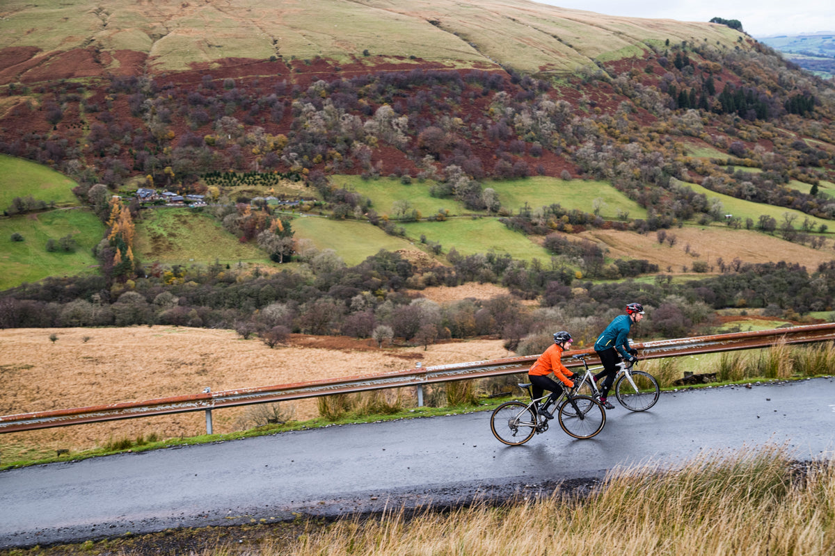 Two cyclists ride along a wet road through a rolling green and brown hills landscape