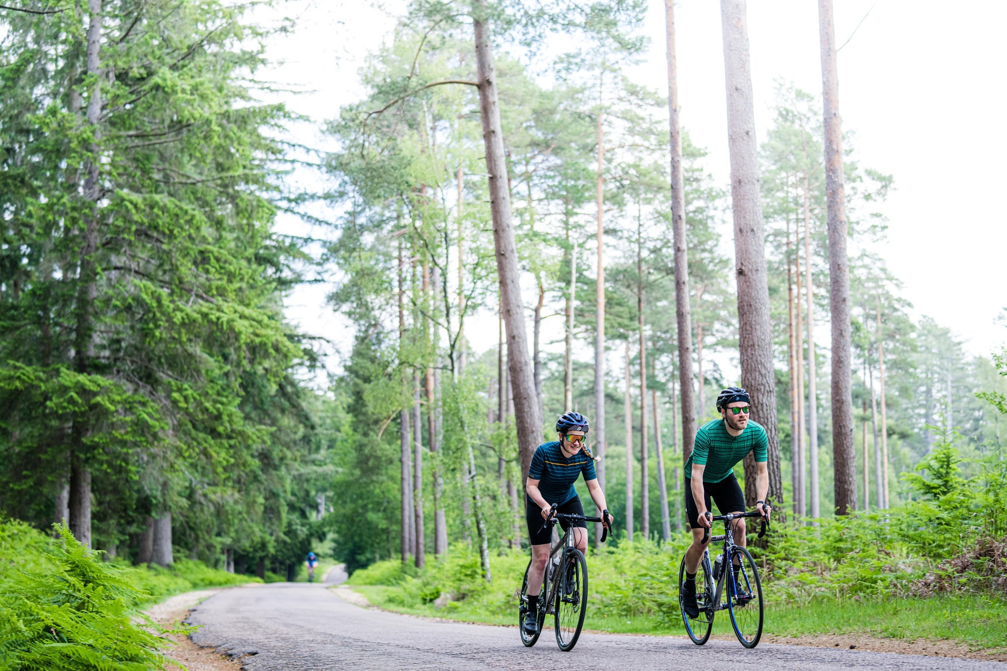 Two cyclists wearing Rivelo cycling apparel, riding on a forest road